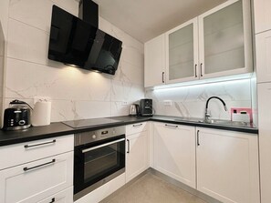 A modern kitchen with white cabinets, a black countertop, a built-in oven, a coffee maker, and a TV mounted above the counter. - Apartment Visnja (Crikvenica)