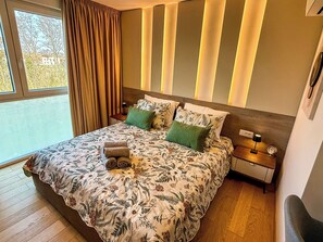 Un dormitorio con una cama grande, una mesita de noche, una lámpara y vistas a árboles por la ventana.