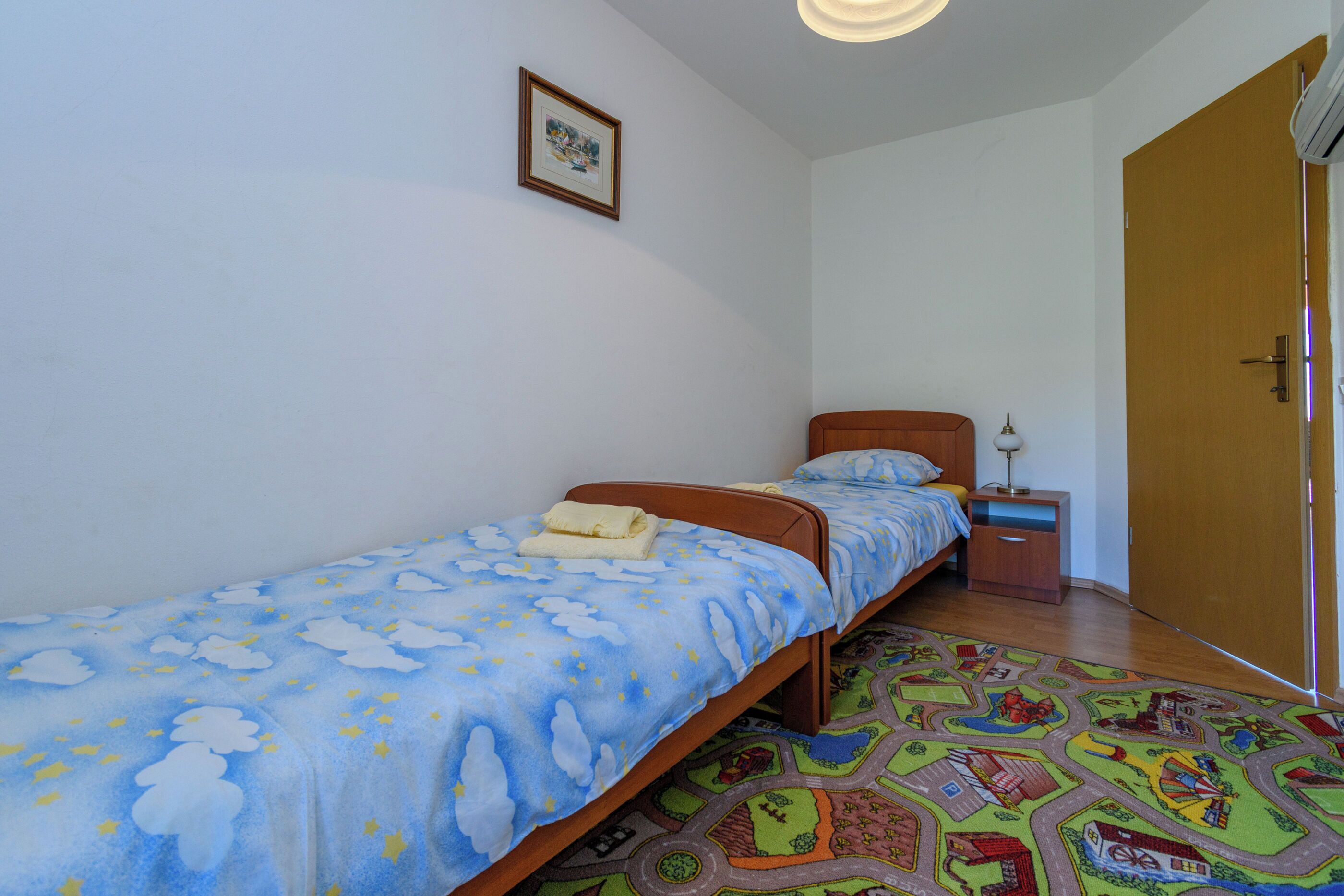 Een eenpersoonsbed met een blauw-witte bedspreide, een klein nachtkastje met een lamp, en een kleurrijk vloerkleed op een houten vloer.