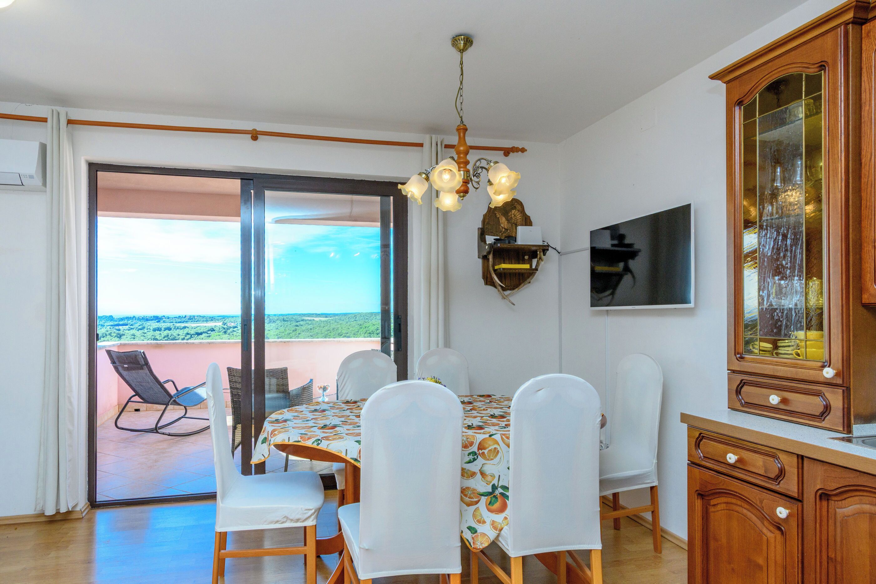 A dining area with a table, chairs, a chandelier, a television, and a view through a glass door.