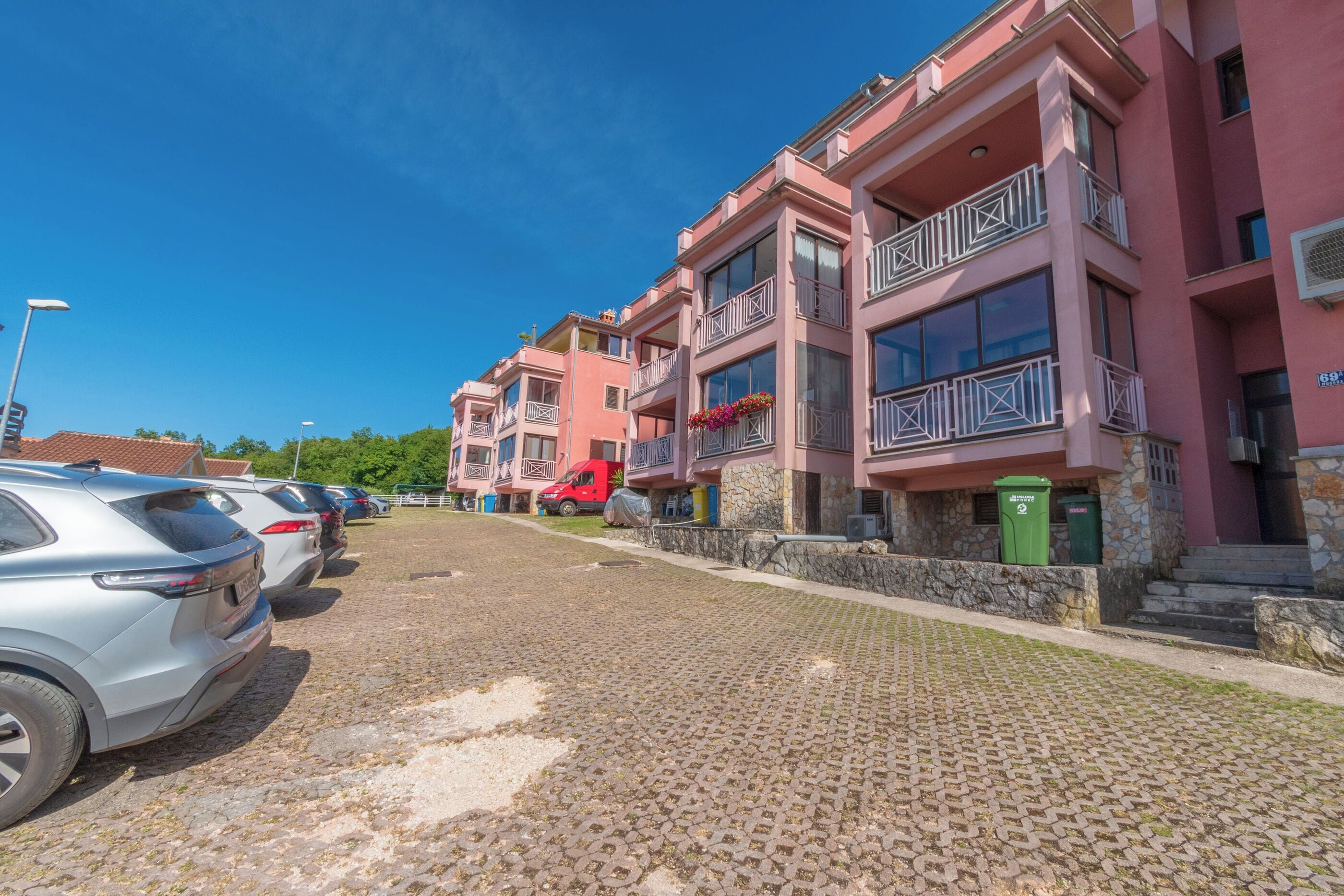 A row of pink buildings with balconies, a cobblestone street, and parked cars.