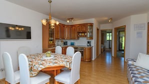 Ein Essbereich mit Tisch, Stühlen und einer Chandelier. Im Hintergrund ist eine Küche mit Holzschränken und Geräten zu sehen.