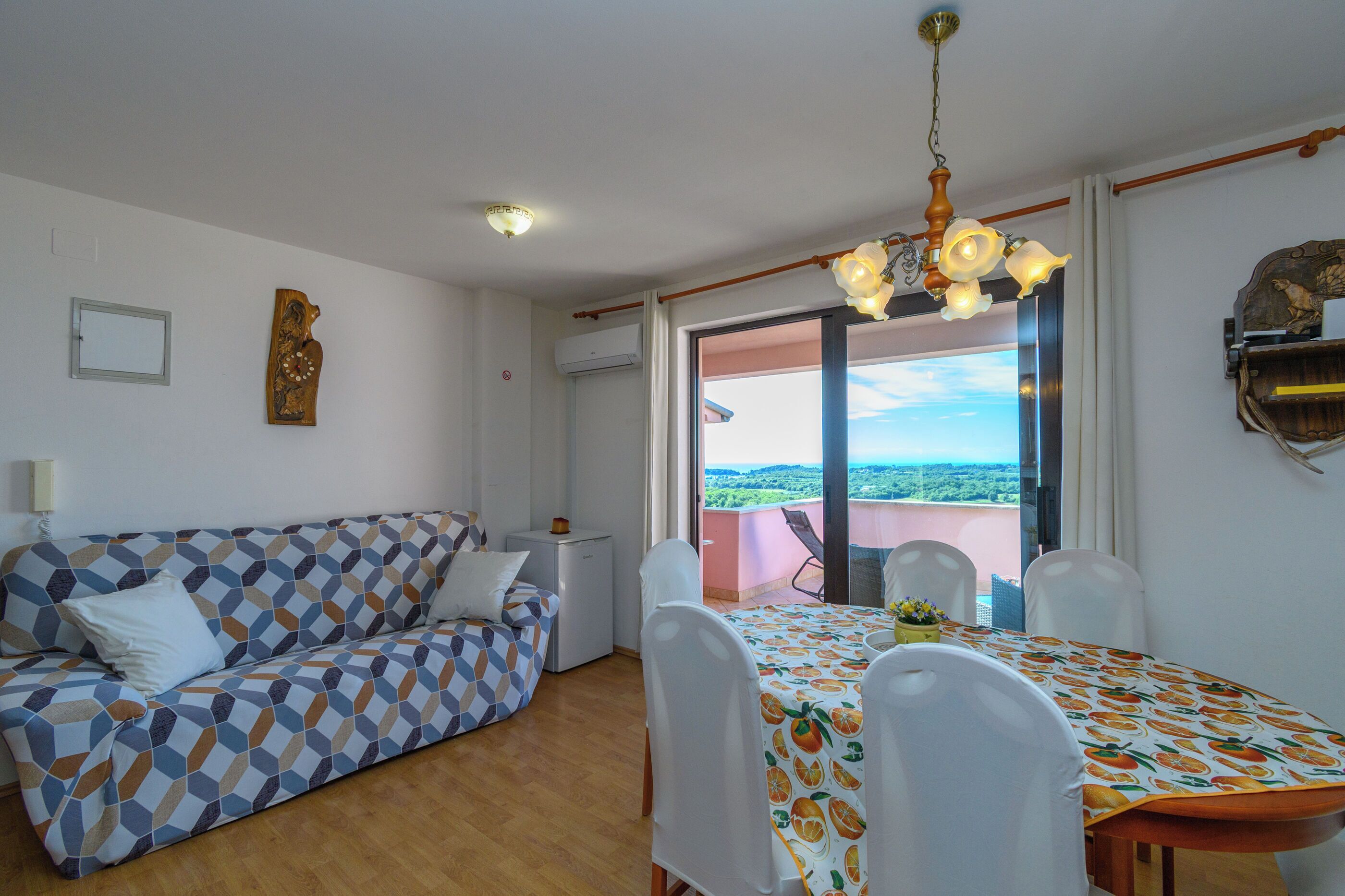 Ein Zimmer mit einer Couch, einem Esstisch mit Stühlen, einer Chandelüster und Blick durch eine Glastür.