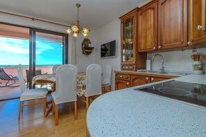 A kitchen with wooden cabinets, a dining area with chairs, a table with a fruit pattern, and a large window. - Apartment Nikola (Porec)