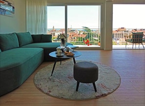 Ein modernes Wohnzimmer mit einem grünen Sofa, einem runden Couchtisch mit Vase und Teekanne sowie Blick auf eine Stadtlandschaft durch große Fenster.