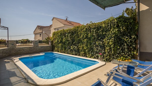 A swimming pool with a tiled deck, a covered seating area, and a hedge.
