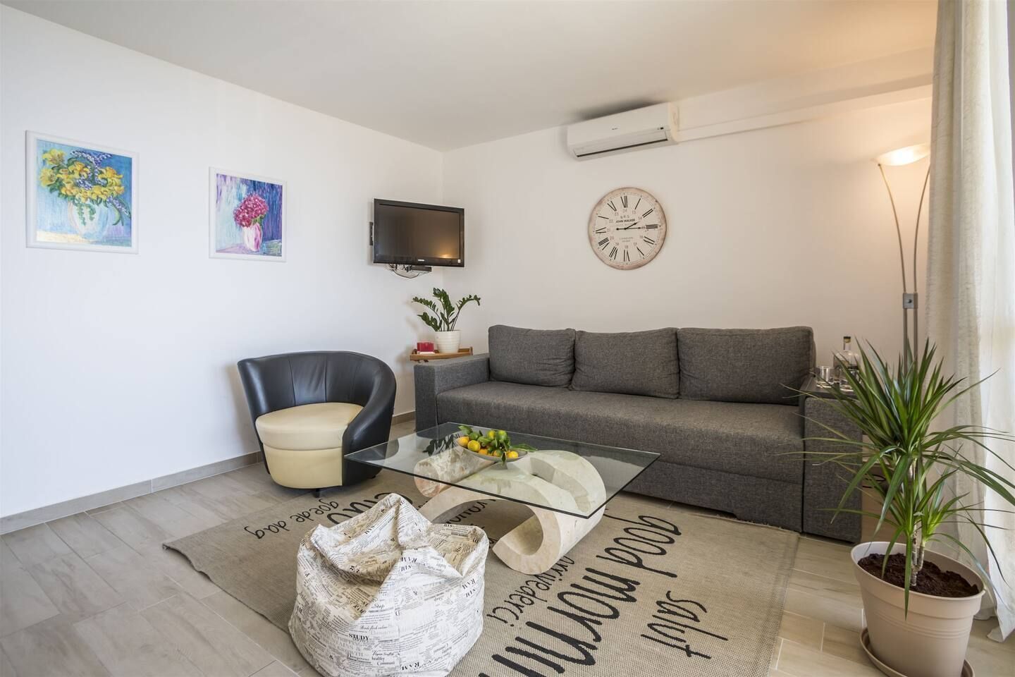Uma sala de estar moderna com um sofá, uma poltrona de couro, uma mesa de centro de vidro, uma televisão de tela plana, um relógio de parede e um vaso com planta.