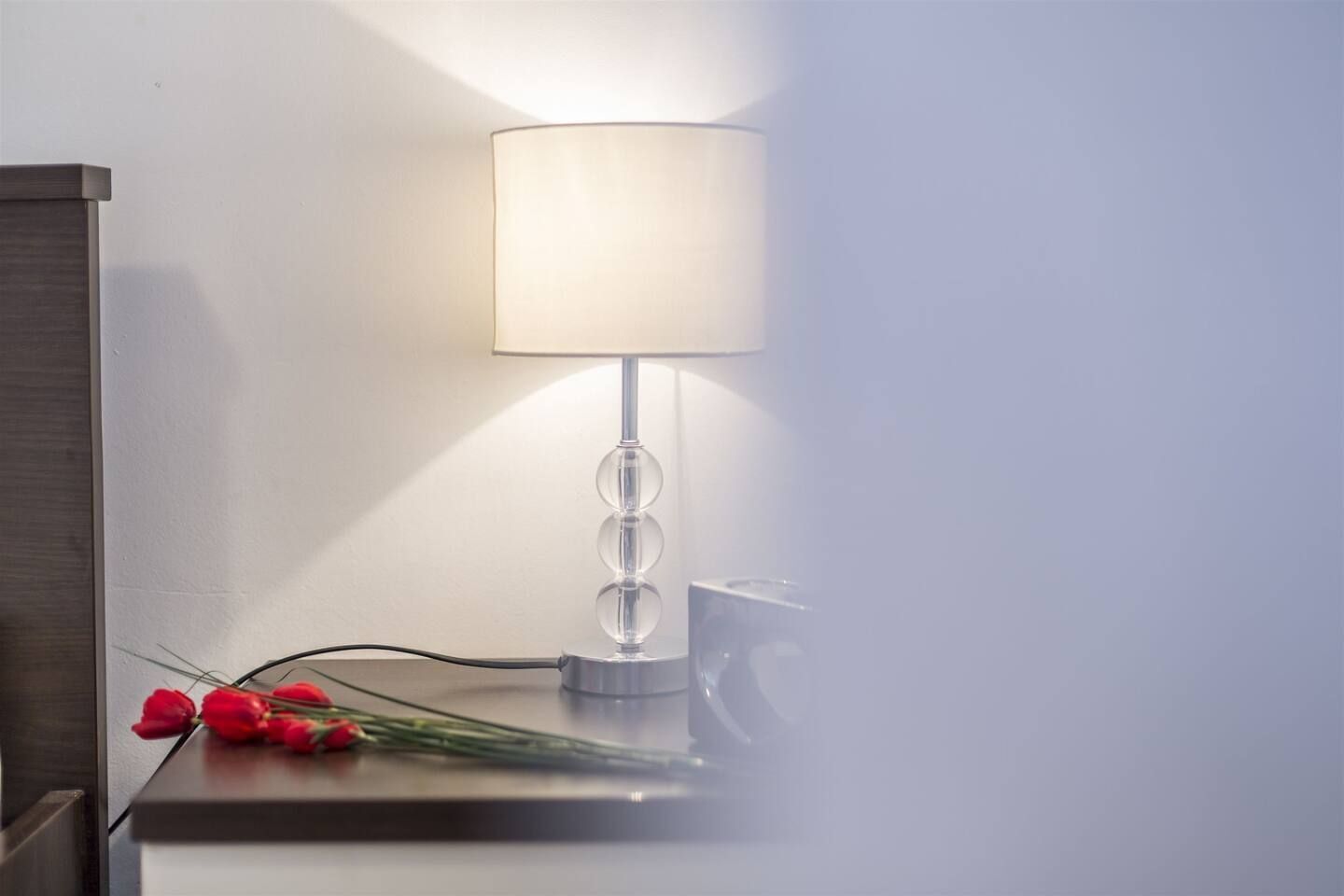 A modern lamp with a clear glass base and a white shade, placed on a wooden surface with a power cord and red tulips beside it.