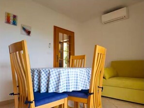 Ein Essbereich mit Tisch, bedeckt mit einer karierten Tischdecke, zwei Holzstühlen mit blauen Kissen, einem gelben Sofa und einer Klimaanlage an der Wand.