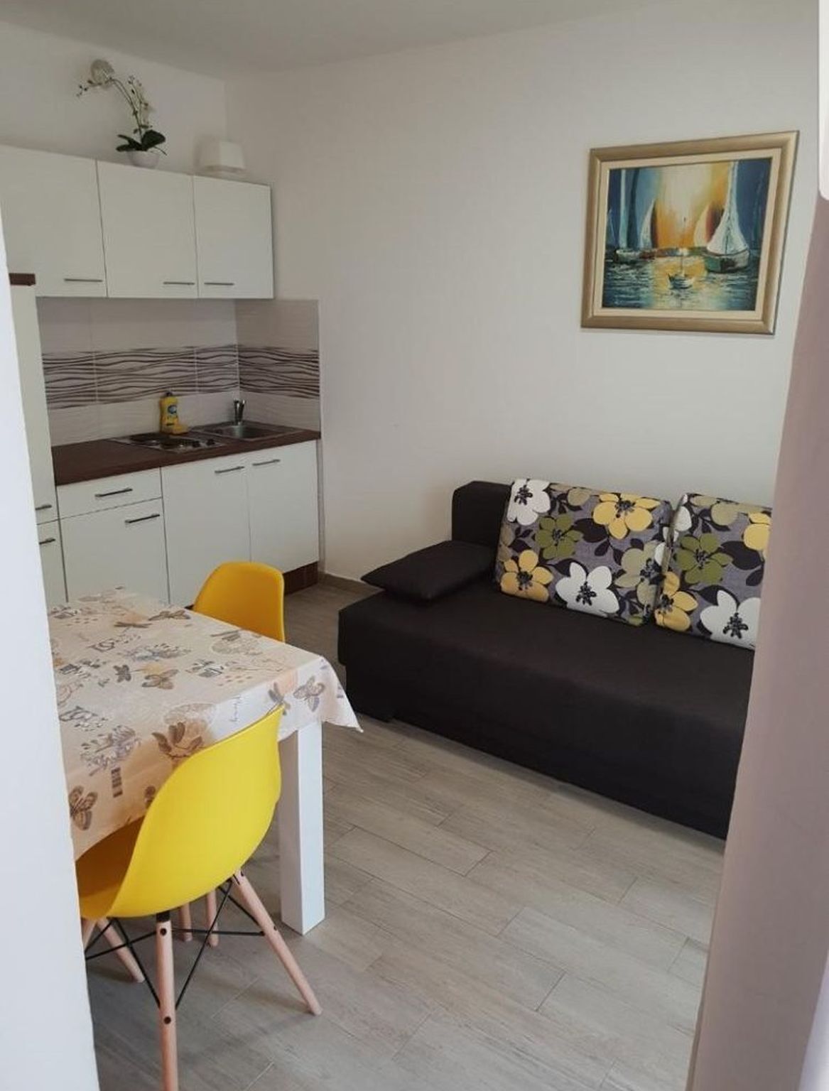 Ein gemütlicher Wohnraum mit einer Küchenzeile, einem Esstisch mit Stühlen, einer Couch mit gemusterten Kissen und einem Bild an der Wand.