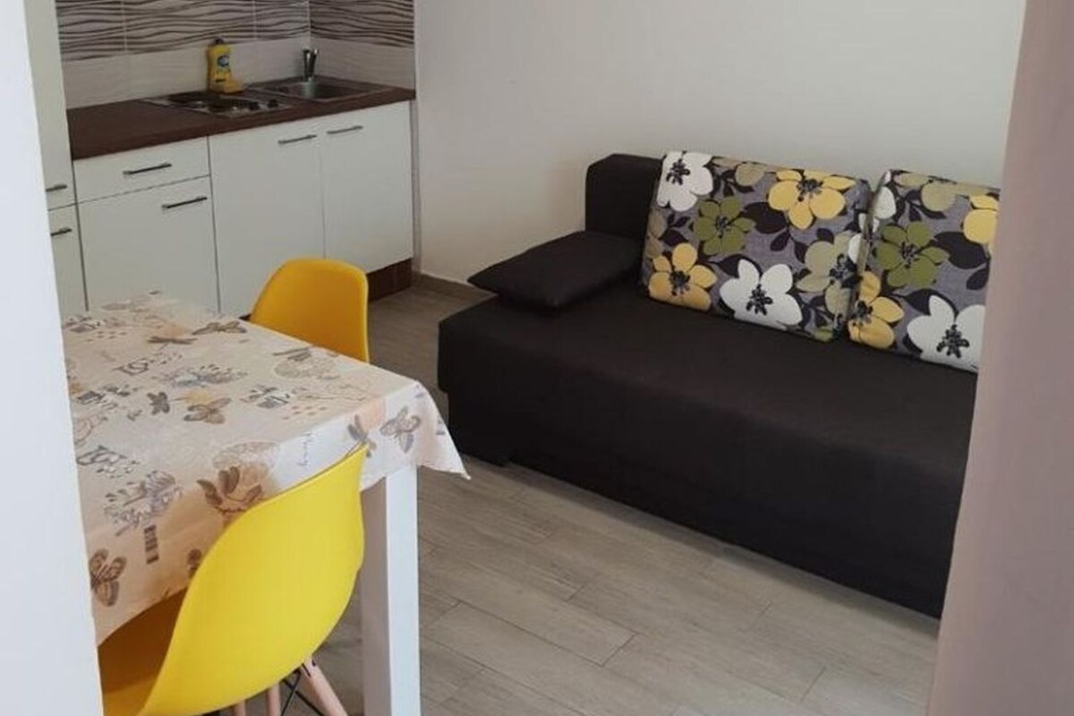 Ein gemütlicher Wohnraum mit einer Küchenzeile, einem Esstisch mit Stühlen, einer Couch mit gemusterten Kissen und einem Bild an der Wand.