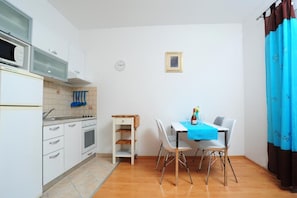Eine kompakte Küche mit weißen Schränken, einer Mikrowelle und einem Spülbecken. Ein Esstisch mit Stühlen und einer blauen Tischdecke steht bereit.