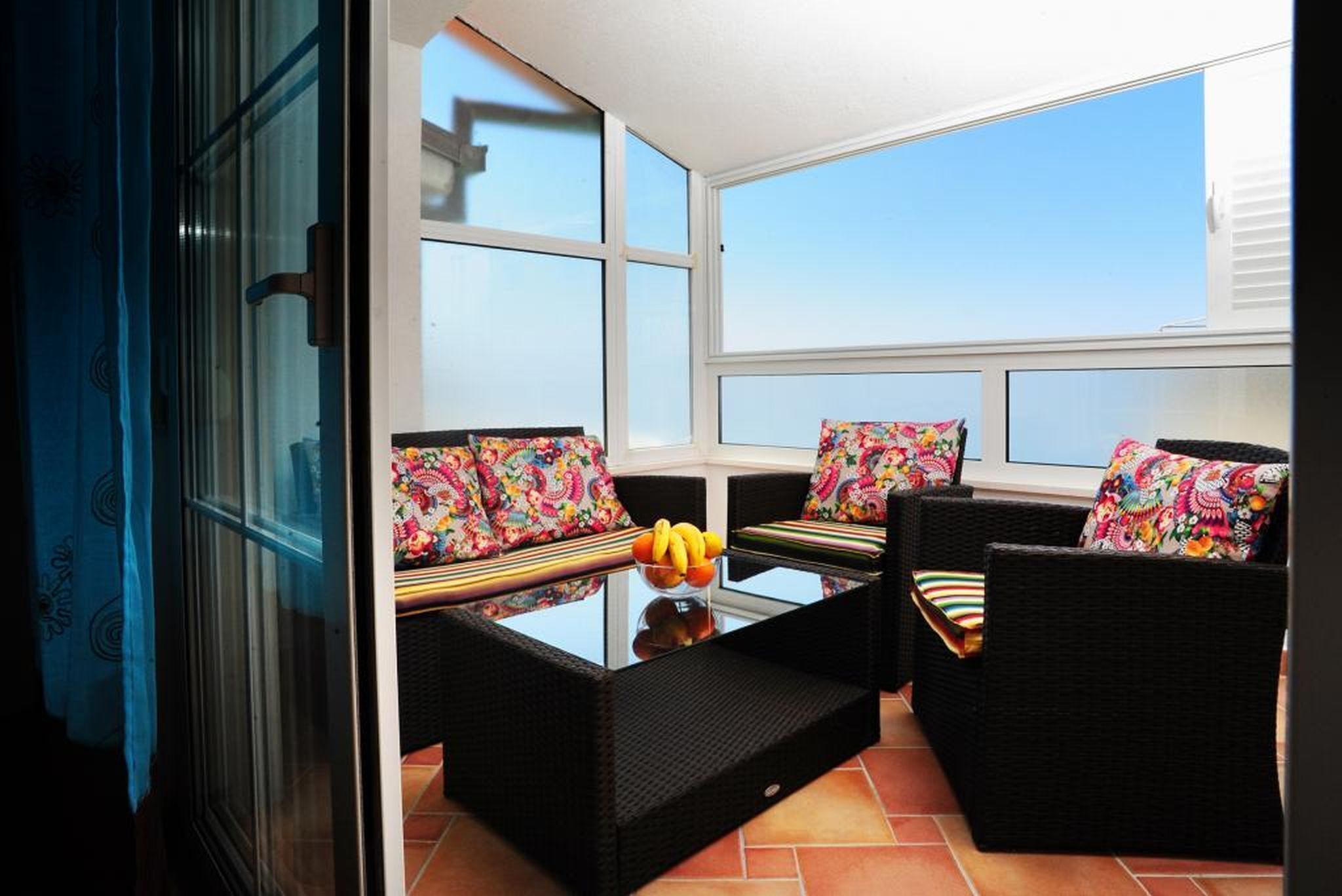 Ein Balkon mit Korbmöbeln und bunten Kissen, ein Glastisch mit Obst und freie Sicht zum Himmel.