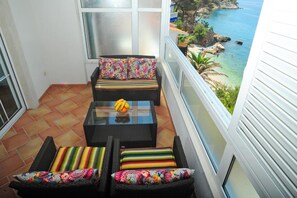 Ein Balkon mit Korbmöbeln, einem Tisch mit Glasplatte und Früchten sowie Blick auf die Küste.