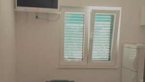 A dining area with a table and chairs, a television mounted on the wall, and a window with blinds. - Apartments Oliva (Sibenik)