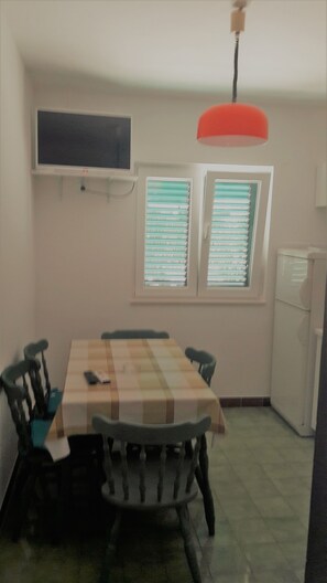 A dining area with a table and chairs, a television mounted on the wall, and a window with blinds. - Apartments Oliva (Sibenik)