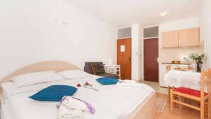 A neatly made bed with pillows, a chair, a small table with a kettle, and a kitchen area with cabinets and a sink. - Apartments Neda (Dugi Rat)