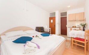 Ein ordentlich bezogenes Bett mit Kissen, ein Sessel, ein kleiner Tisch mit Wasserkocher und eine Küchenzeile mit Schränken und Spüle.
