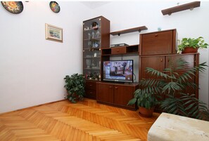 Ein Wohnzimmer mit Holzdielen im Fischgrätmuster, einem Fernseher, Holzschränken und Topfpflanzen.