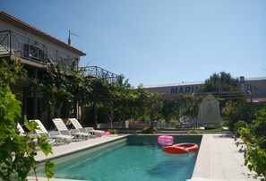Ein Schwimmbecken mit Liegestühlen und aufblasbaren Ringen, umgeben von Grünflächen und einem Gebäude mit der Beschriftung 'MARINA'.