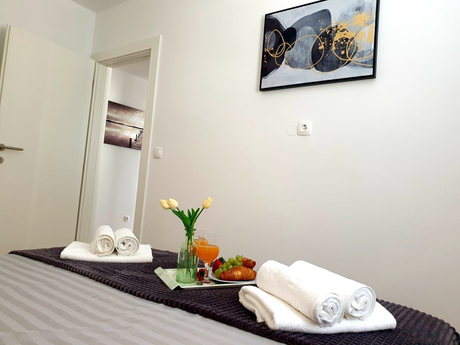 Quarto de hotel com cama, quadro, vaso com flores, prato de frutas e toalhas.