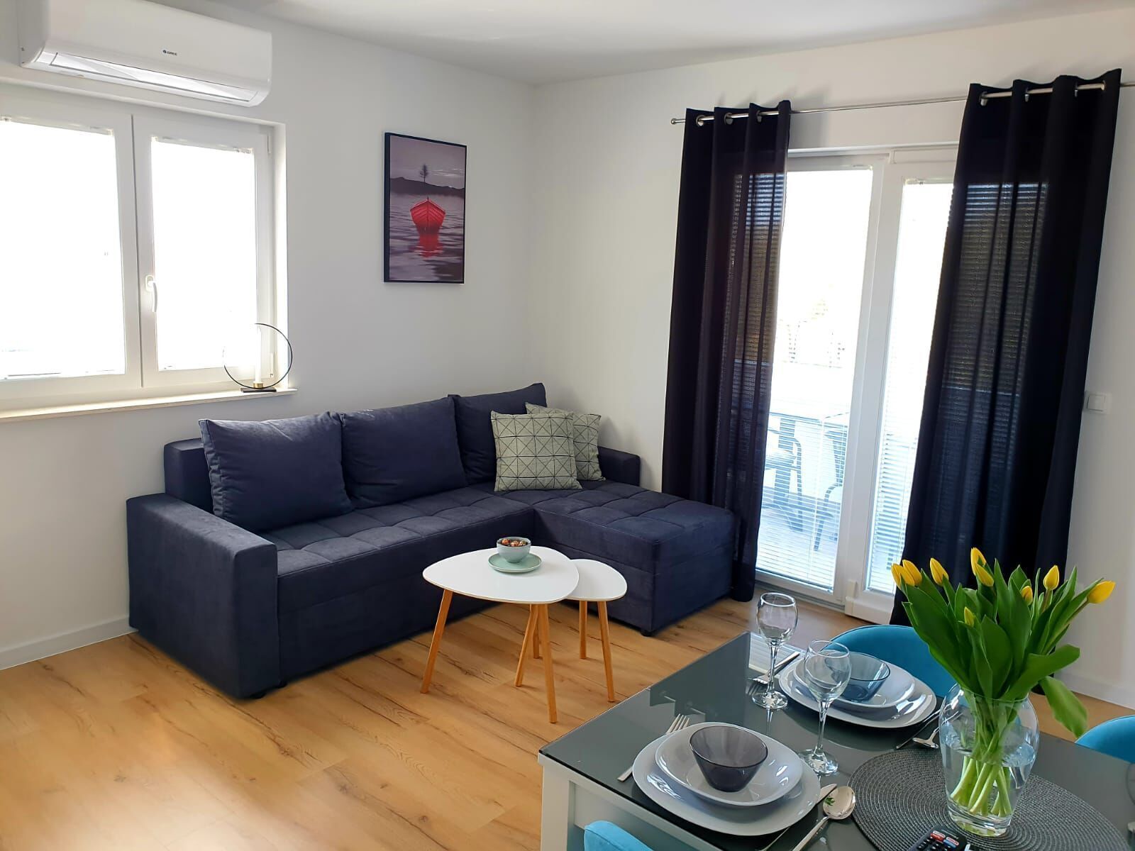 Un salon moderne avec un canapé bleu foncé, une petite table blanche, une table à manger avec des chaises et une fenêtre avec des rideaux.
