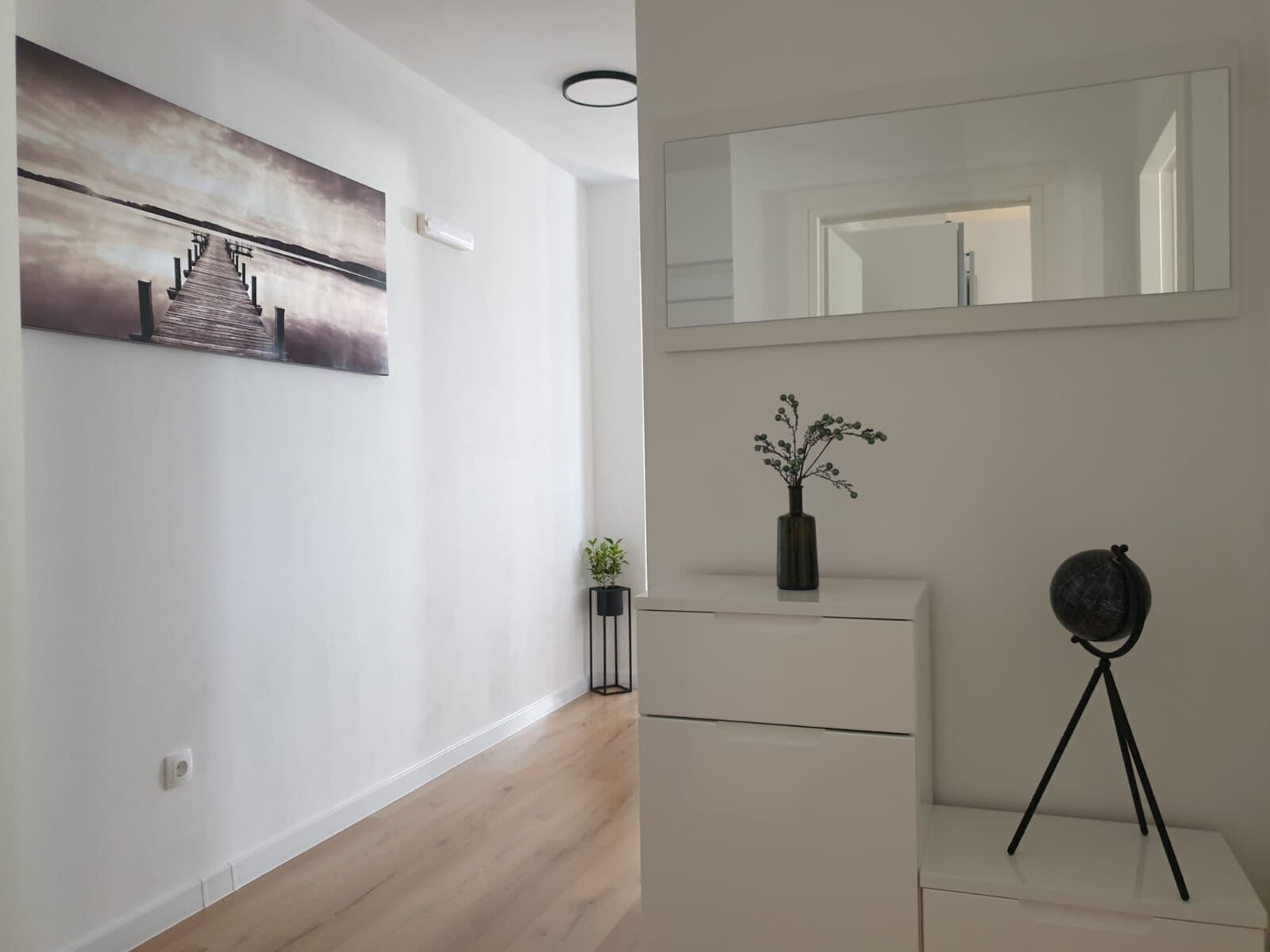 Ein modernes Interieur mit Holzboden, einer weißen Wand mit gerahmtem Bild, einer kleinen Pflanze, einer Vase mit Blumen, einem Spiegel und einem Stativ.