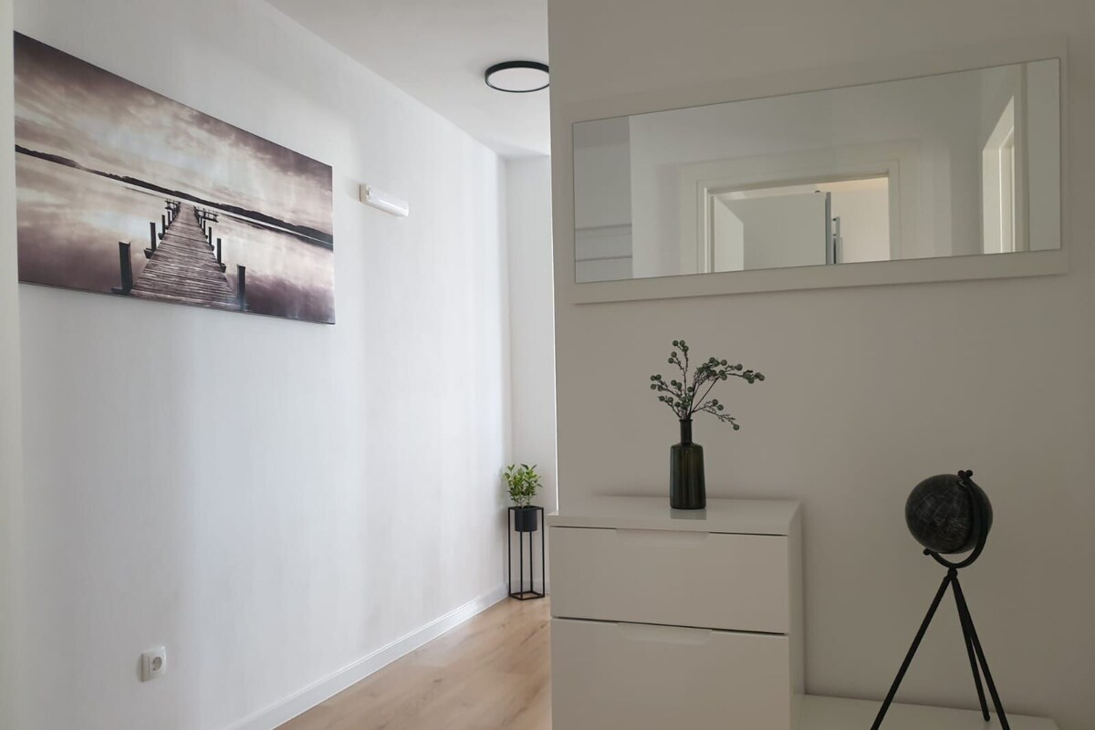 A modern interior with a wooden floor, a white wall with a framed picture, a small plant, a vase with flowers, a mirror, and a tripod.