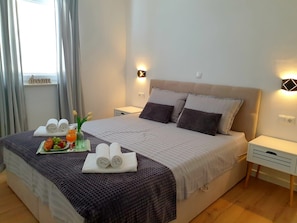 Ein ordentlich bezogenes Bett mit Kissen, ein Nachttisch, ein Fenster mit Vorhängen und ein Tablett mit Handtüchern und Obst.