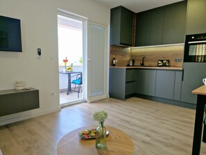 A modern kitchen with dark cabinets, a built-in oven, and a dining area with a round table and chairs. - Apartment Tea (Vodice)