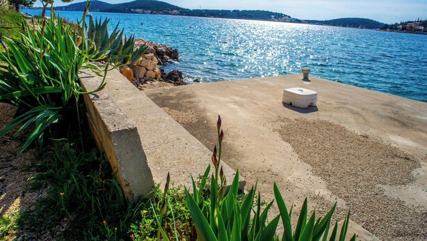 A concrete waterfront area with a stone wall, a small white structure, and green vegetation. - Apartment Kaleta (Rogoznica)