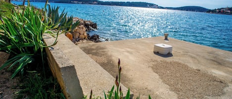 Un área costera de concreto con un muro de piedra, una pequeña estructura blanca y vegetación verde.