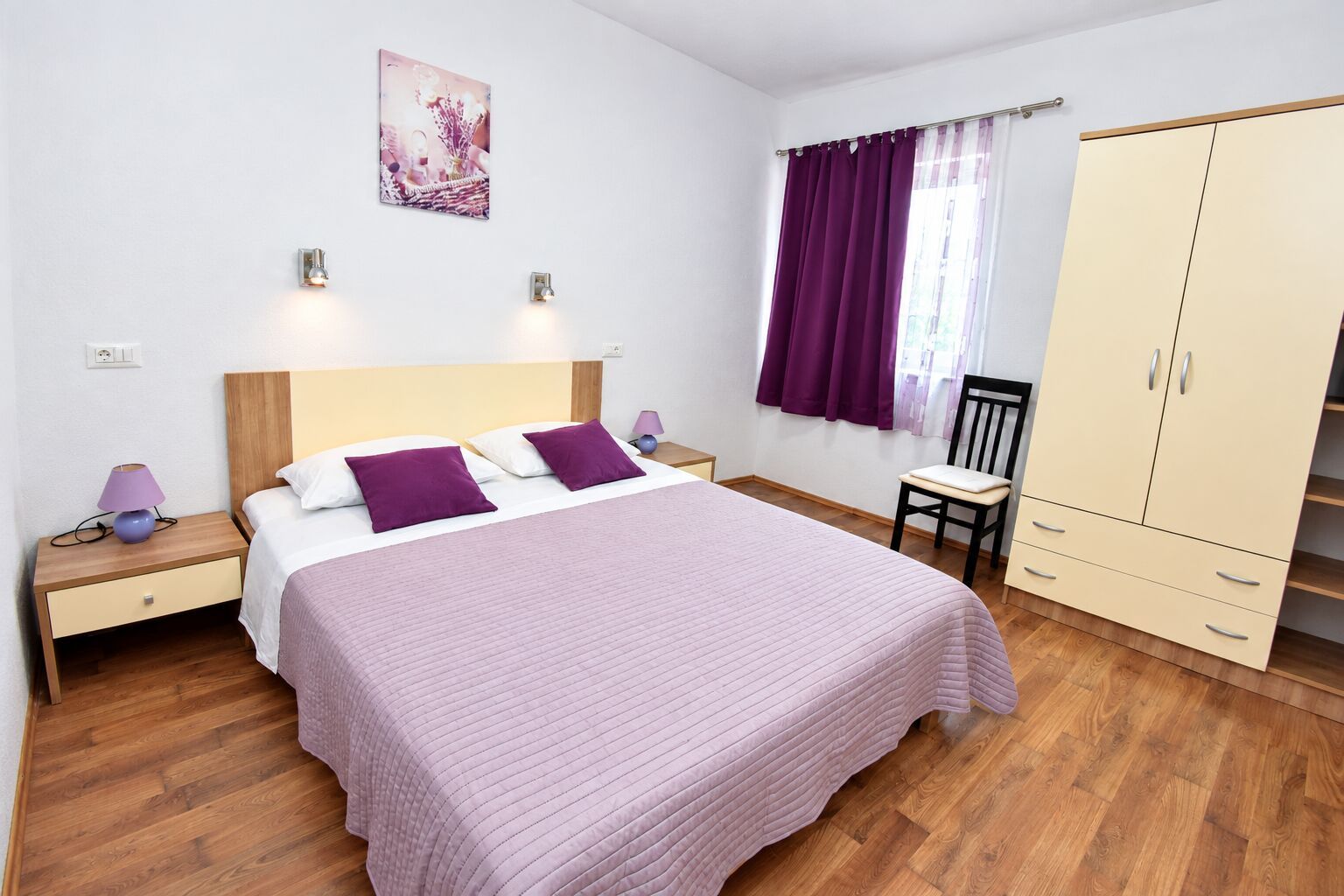 A bedroom with a bed, nightstands, a chair, a wardrobe, and a window with curtains.