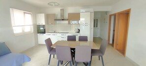 A compact kitchen with white cabinets, a dining table with four chairs, and a blue sofa in the adjacent room. - Apartment Kaleta (Rogoznica)