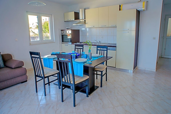 A modern kitchen with a dining area, featuring a table set for four, chairs, a sofa, and a window with a view. - Apartment Kaleta (Rogoznica)