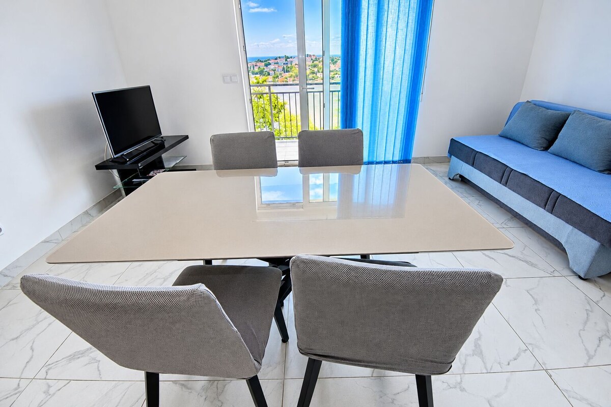 A modern living room with a large table, two grey chairs, a sofa, and a TV.
