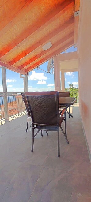 A covered outdoor patio with a table and chairs, large windows, and a view of the sky and distant landscape. - Apartment Kaleta (Rogoznica)