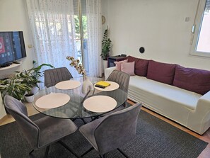 Ein modernes Wohnzimmer mit einem Glastisch, grauen Stühlen, einem weißen Sofa mit roten Kissen und einer Zimmerpflanze.