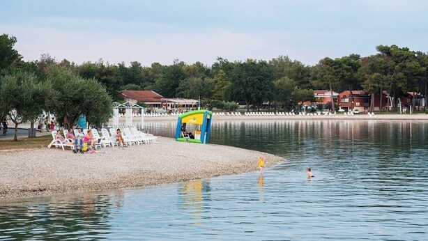 A lakeside area with a small inflatable play structure, a row of beach chairs, and a building in the background. - Apartments Durdica (Umag)