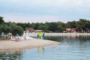 Ein Uferbereich mit einer kleinen aufblasbaren Spielanlage, einer Reihe Strandstühle und einem Gebäude im Hintergrund.