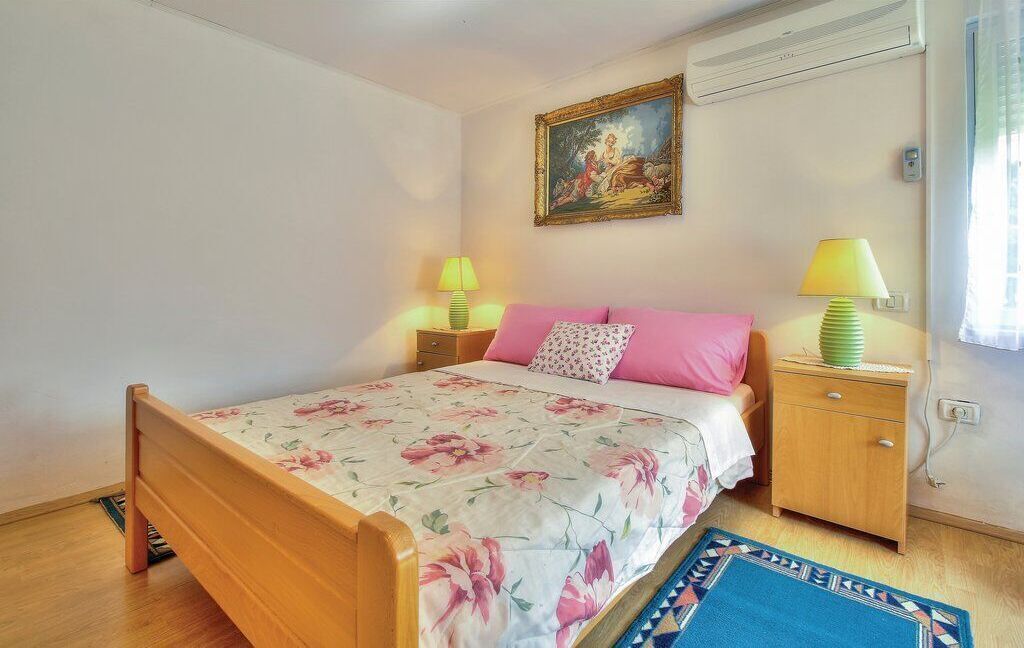Un dormitorio con una cama de madera, ropa de cama estampada con motivos florales, un cojín rosa, una mesita de noche con lámpara, un cuadro en la pared y una ventana con cortinas.