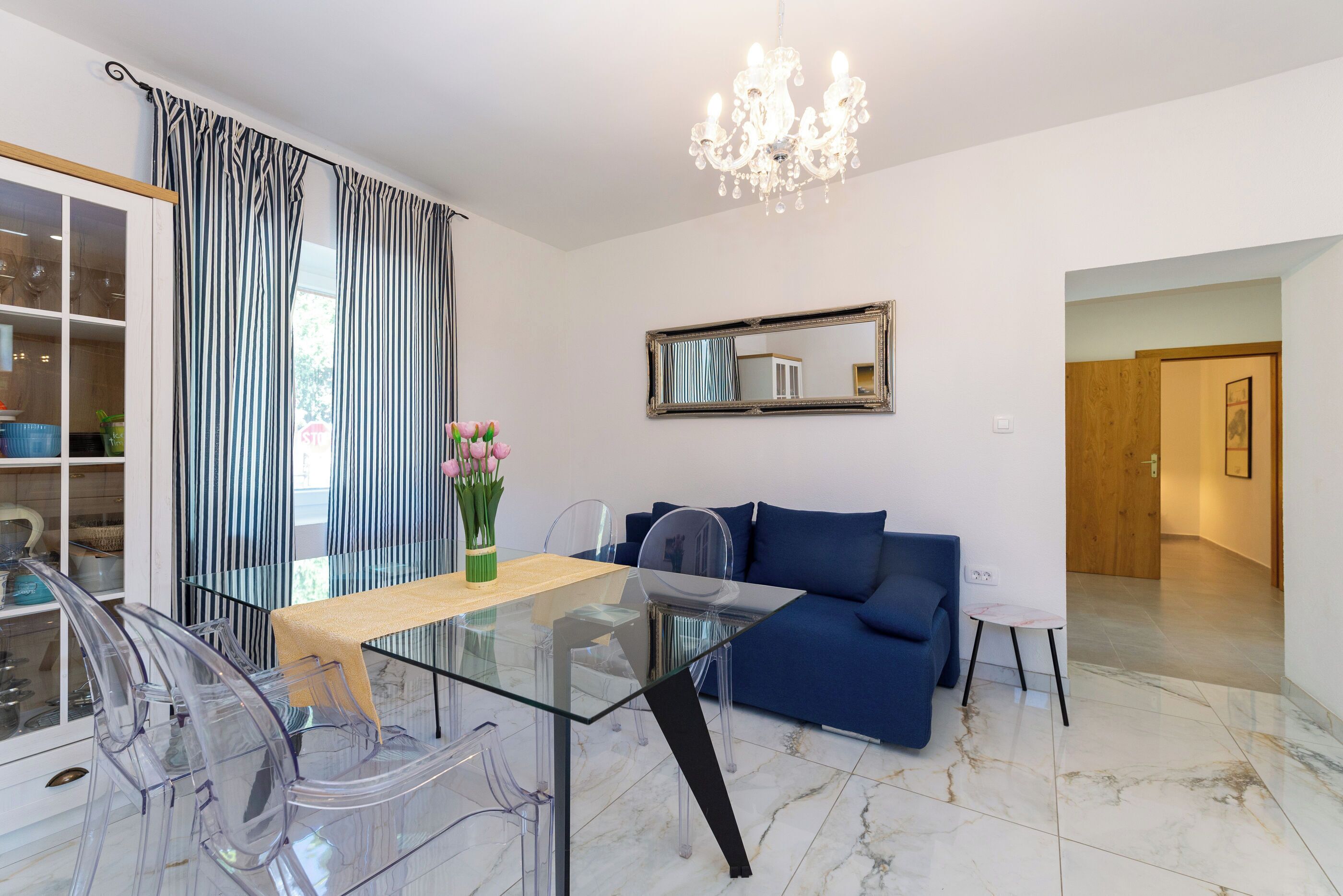 A modern living room with a glass dining table, clear plastic chairs, a blue sofa, a chandelier, and a large mirror on the wall.