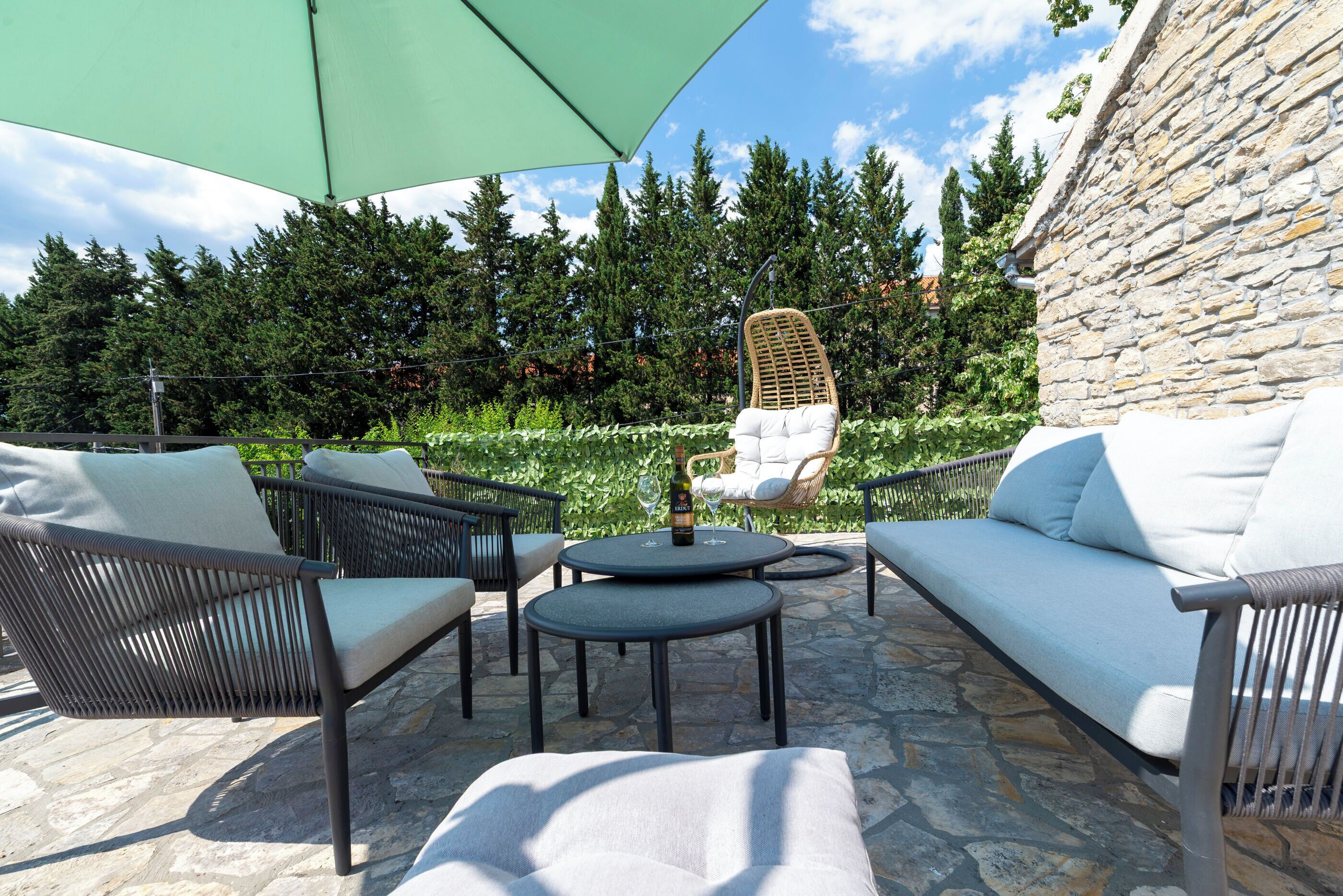 An outdoor patio with wicker furniture, a stone floor, and a view of trees and a stone wall.