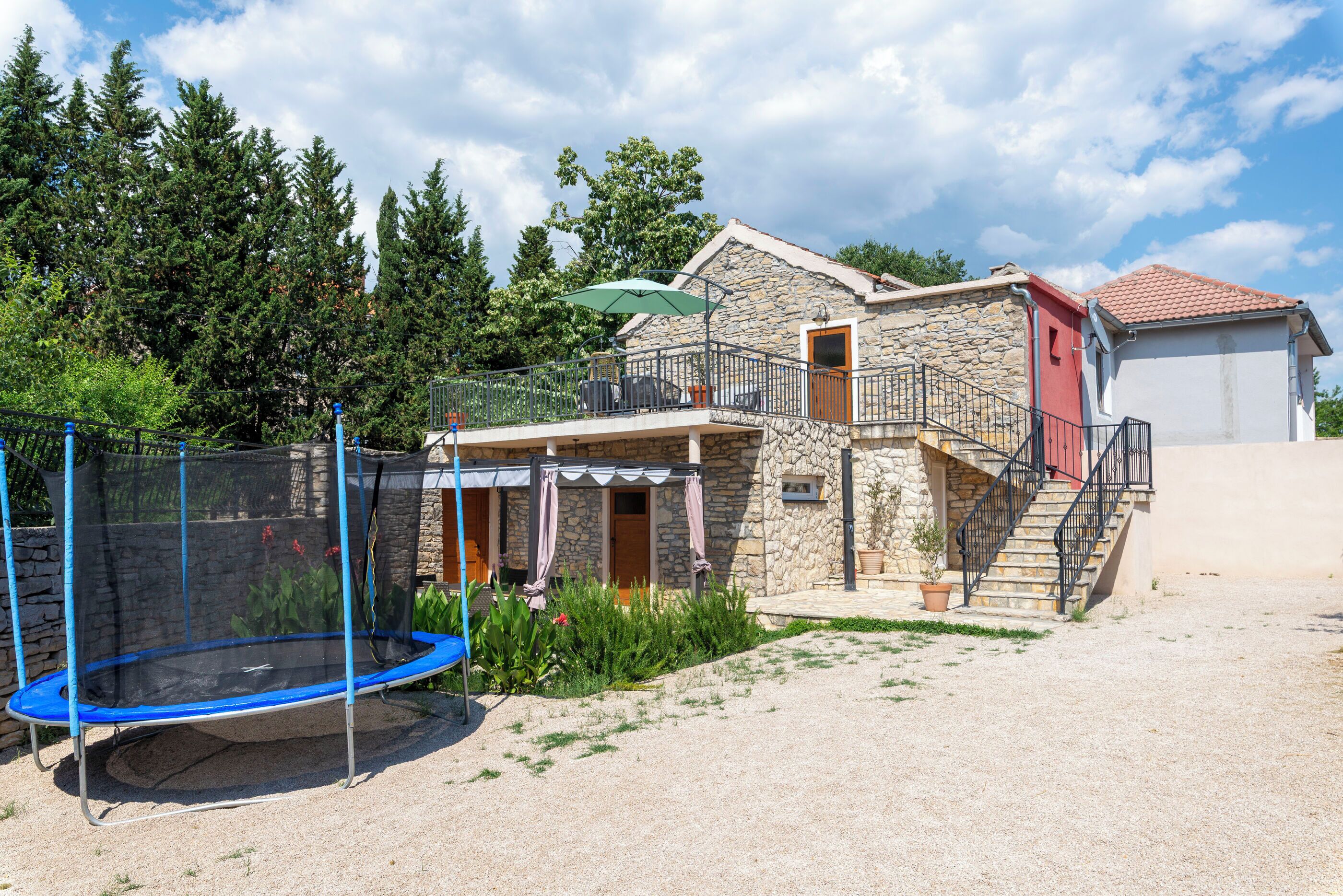 Une maison en pierre avec un balcon, un trampoline et un espace extérieur aménagé avec une zone de repos couverte.