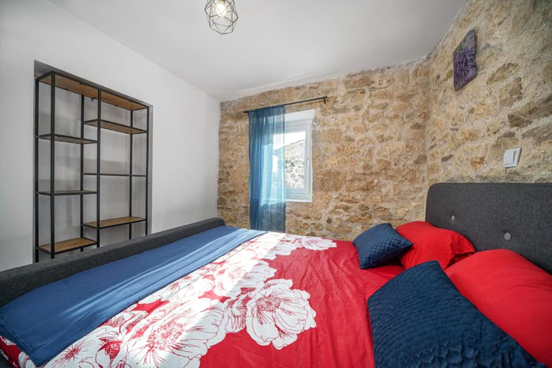 Un dormitorio con pared de piedra, una cama con ropa de cama roja y azul, una estantería de madera y una ventana con cortinas azules.