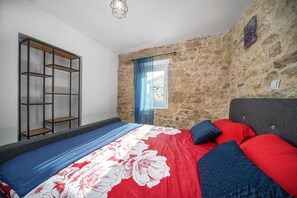 Ein Schlafzimmer mit Steinwand, einem Bett mit rot-blauer Bettwäsche, einer Holzregaleinrichtung und einem Fenster mit blauen Vorhängen.