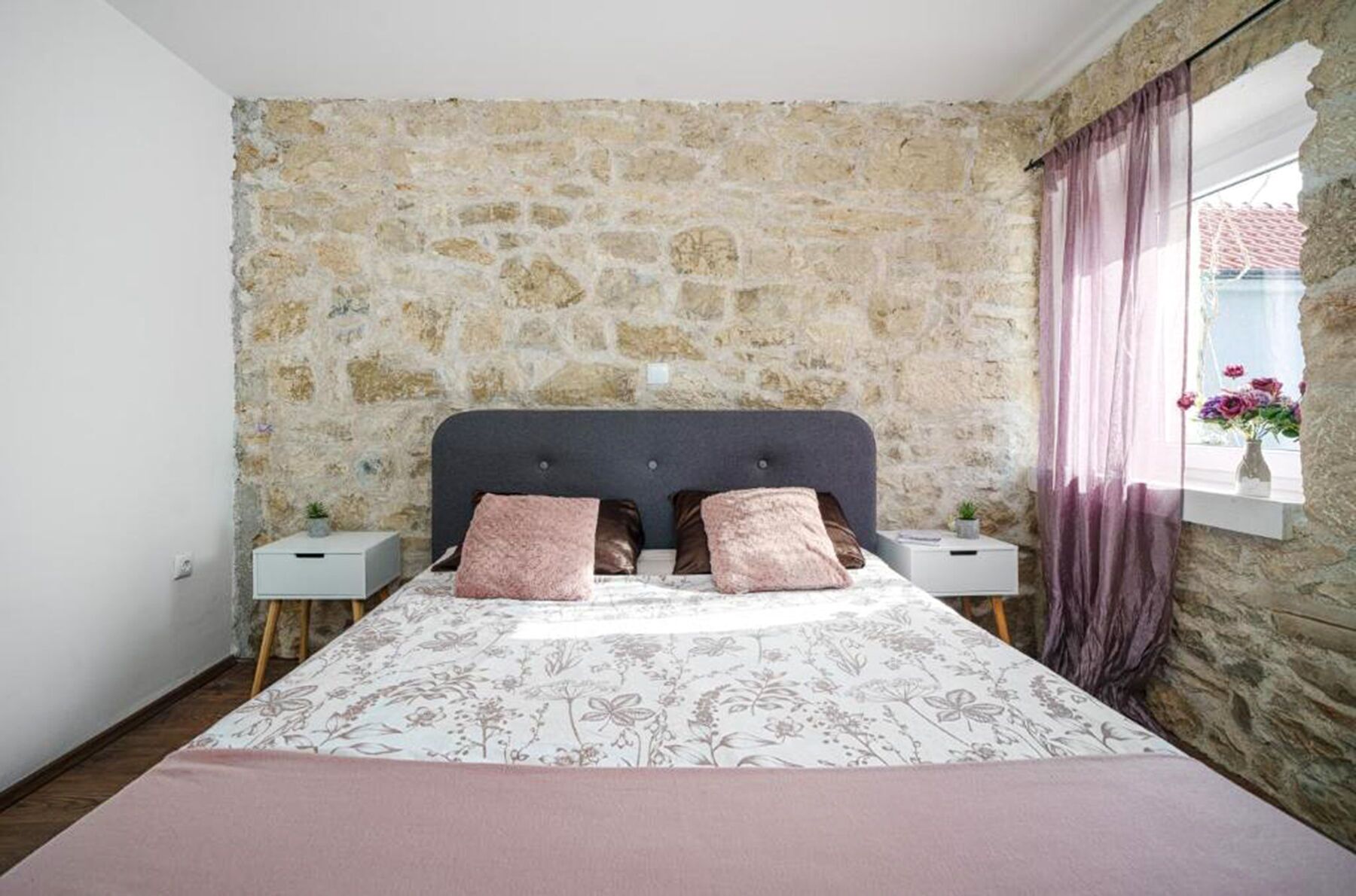 A bedroom with a stone wall, a bed with floral bedding, two nightstands, and a window with curtains.