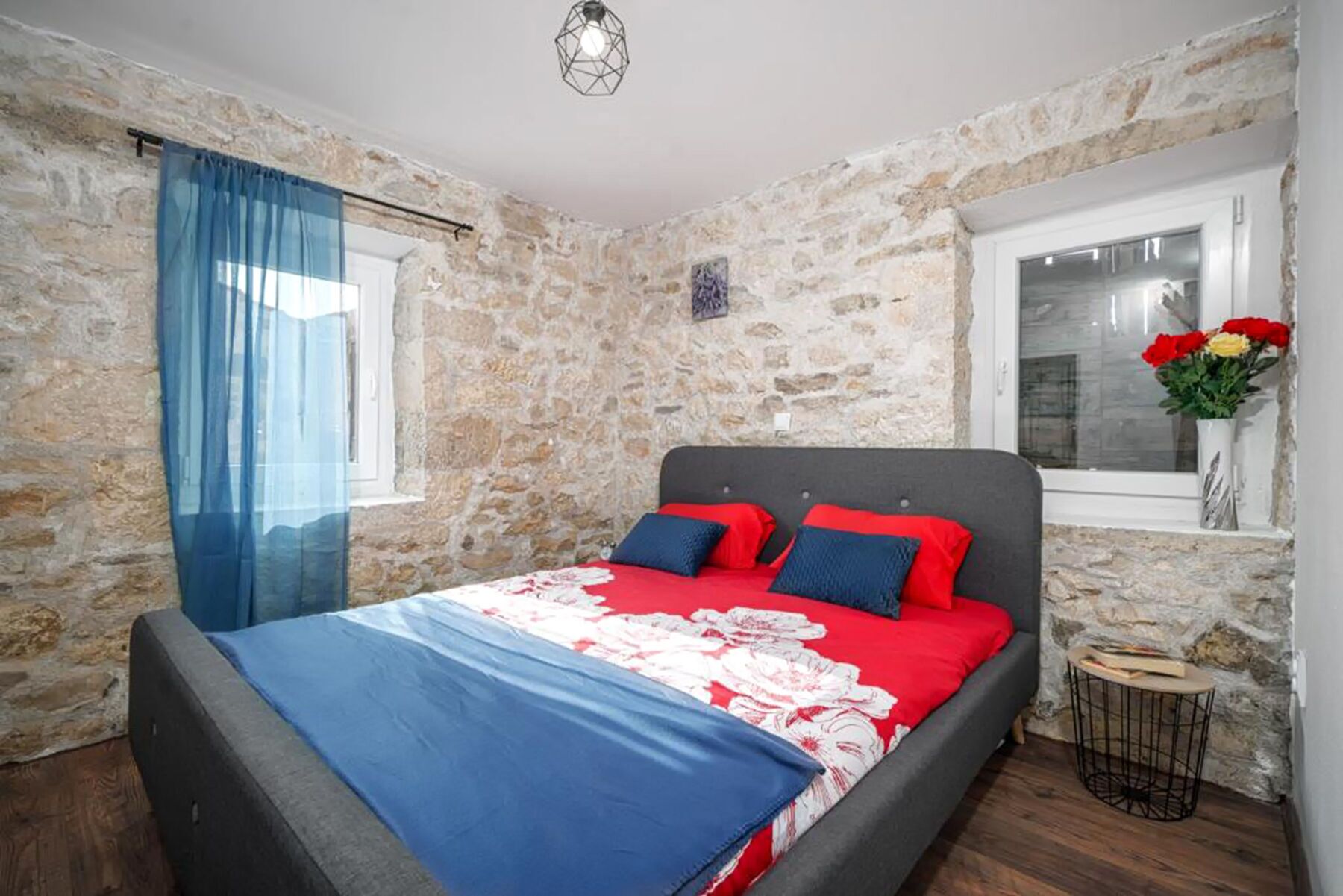 A bedroom with stone walls, a bed with red and blue bedding, a window with a view, and a small table with a vase of flowers.