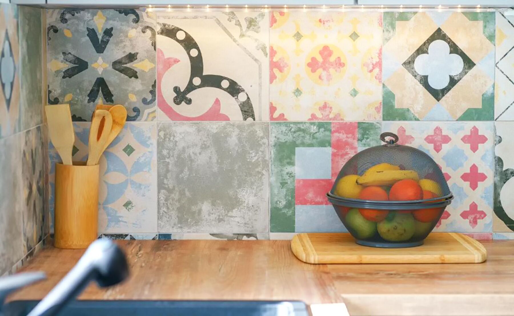 Une cuisine avec des carreaux de céramique aux couleurs vives, une planche à découper en bois, un saladier rempli de fruits et un porte-ustensiles.