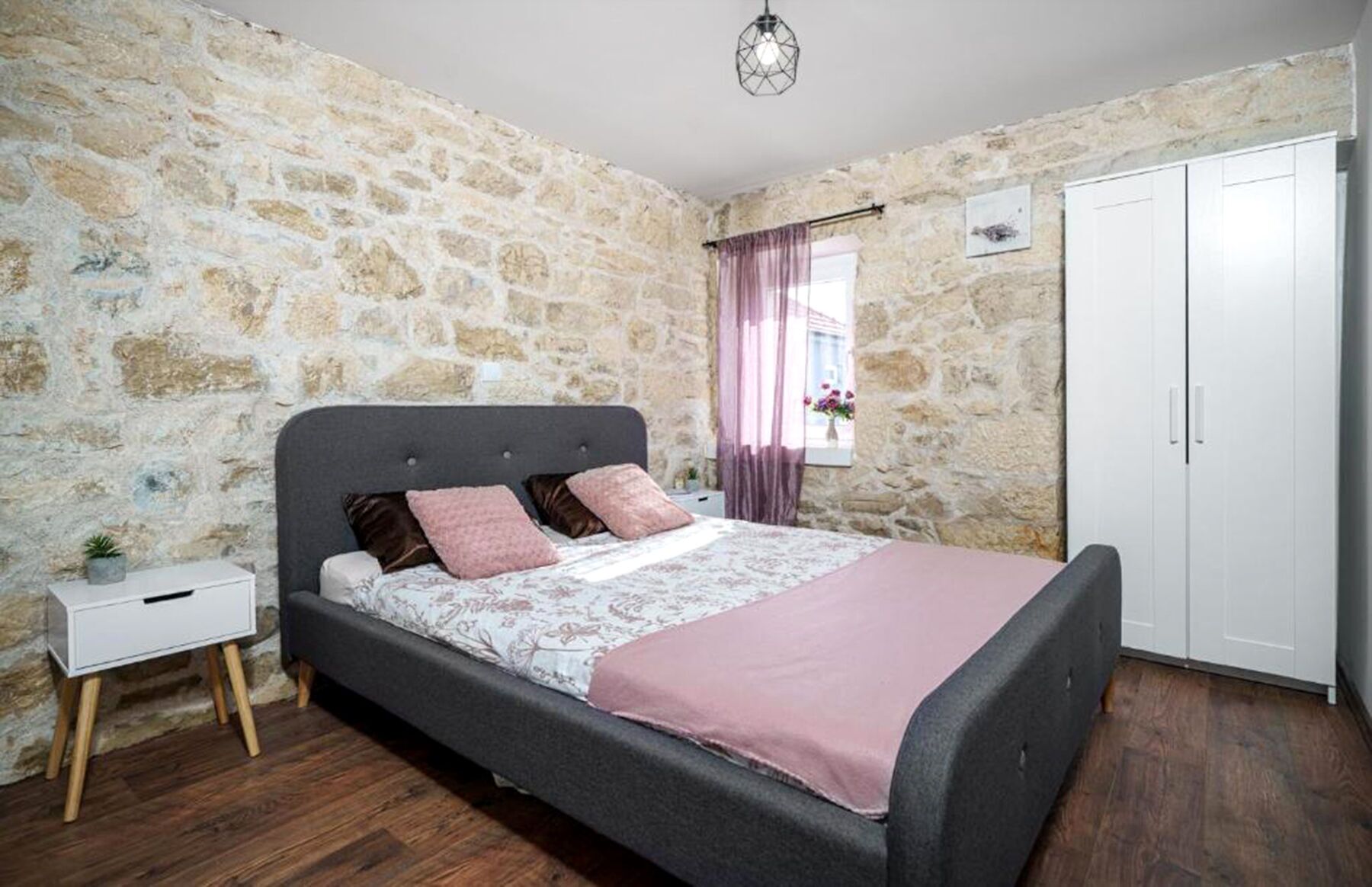 A bedroom with a stone wall, a bed with a grey headboard, a white bedside table, and a wardrobe.